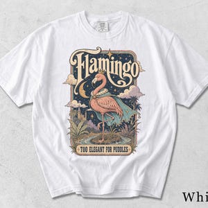Pode incluir: Uma t-shirt branca de manga curta com uma estampa gráfica de um flamingo usando uma coroa e uma capa. O design inclui uma lua crescente, estrelas, nuvens e palmeiras, com o texto Flamingo e Too Elegant For Puddles em uma fonte vintage.