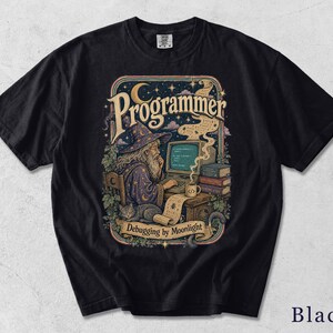 Może przedstawiać: Czarny t-shirt z nadrukiem graficznym przedstawiającym czarodzieja siedzącego przy komputerze z kotem, książkami i kubkiem kawy. Napis "Programmer" znajduje się u góry, a "Debugging by Moonlight" u dołu, w otoczeniu gwiazd i roślinności.