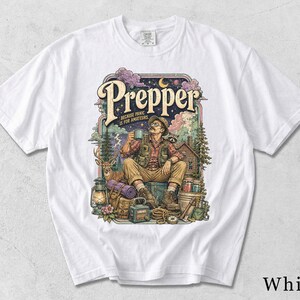 Könnte beinhalten: Ein weißes kurzärmeliges T-Shirt mit einem farbenfrohen Grafikdruck, der eine Person im Freien mit Campingausrüstung, einem Hirsch und einem Waschbären zeigt. Der Text lautet Prepper und Because panic is for amateurs vor einem Sternenhimmel.