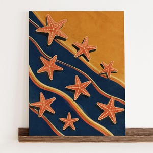 Könnte beinhalten: Ein gerahmter Wandkunstdruck mit einer Reihe orangefarbener Seesterne vor einem Hintergrund aus wellenförmigen, geschichteten Bändern in Dunkelblau, hellem Orange und Creme, eingefasst in einen dünnen, goldfarbenen rechteckigen Rahmen.