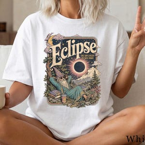 Könnte beinhalten: Ein weißes T-Shirt mit einem Grafikdruck eines Zauberers, der während einer Sonnenfinsternis unter einem Baum liegt. Der Text lautet Eclipse und The sun called in sick today. Die Illustration zeigt eine Mondsichel, Wolken, Bücher und einen Becher.