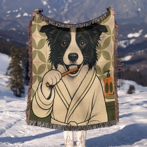 May include: A woven throw blanket featuring a black and white Border Collie wearing a beige bathrobe and brushing its teeth with a wooden toothbrush. The background has a green geometric pattern and an orange soap dispenser on a counter.
