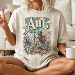 Könnte beinhalten: Ein elfenbeinfarbenes T-Shirt mit einer grafischen Illustration einer Ameise, die mit Rucksack und Wanderstock wandert. Der Text lautet Ant, Small Guy, Huge Plans. Das Design zeigt Berge, Bäume, Pilze, Blumen und eine Sonne im Retro-Stil.