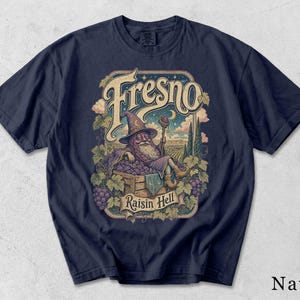 Könnte beinhalten: Ein marineblaues T-Shirt mit einer grafischen Illustration eines Zauberers, der in einer Holzkiste voller violetter Trauben sitzt. Der Text "Fresno" steht in großer Schrift darüber und "Raisin Hell" auf einem Banner darunter.