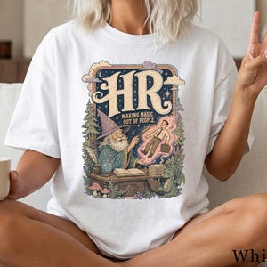 Könnte beinhalten: Ein weißes T-Shirt mit einer grafischen Illustration eines Zauberers an einem Schreibtisch mit Kristallkugel, Büchern und einem kleinen Drachen. Der Text lautet HR MAKING MAGIC OUT OF PEOPLE. Das Design zeigt eine schwebende Person im Anzug.