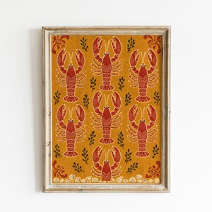 Könnte beinhalten: Ein gerahmter, rechteckiger Wandkunstdruck mit einem sich wiederholenden Muster aus roten Hummern auf senfgelbem Hintergrund. Das Design enthält kleine dunkelgrüne Zweige zwischen den Hummern und dekorative rote Schnörkel in den Ecken.