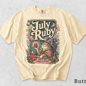 Könnte beinhalten: Ein butterfarbenes T-Shirt mit einer Grafik einer Katze, die einen Zaubererhut und ein Gewand trägt und einen leuchtend roten Kristall hält. Der Text lautet July Ruby und Born to shine, refuses to be subtle, umgeben von bunten Wolken, Sternen und Blättern.