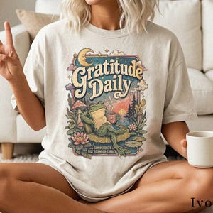 May include: An ivory t-shirt featuring a graphic print of a frog sitting on a lily pad, holding a mug. The design includes a crescent moon, stars, mushrooms, and the text "Gratitude Daily" and "Complaints Use Too Much Energy" in a colorful frame.