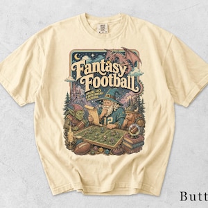 May include: A cream-colored t-shirt featuring a graphic print with the text Fantasy Football, Brains, Luck & Questionable Decisions. The illustration depicts a wizard, a goblin, and a viking playing a board game with a dragon overhead.
