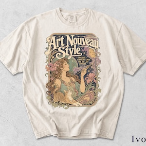 Può includere: Una t-shirt color avorio con una stampa grafica raffigurante una persona che tiene in mano una penna, un gatto e il testo "Art Nouveau Style" e "Because straight lines are so last century." Il design include elementi floreali, una luna e motivi.