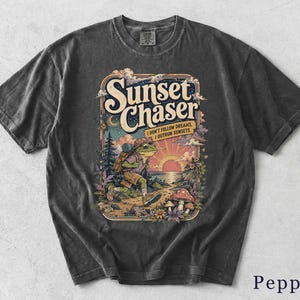 Könnte beinhalten: Ein anthrazitfarbenes Kurzarm-T-Shirt mit einem Grafikdruck eines wandernden Frosches mit Rucksack. Der Text lautet Sunset Chaser und I Don't Follow Dreams, I Outrun Sunsets. Die Illustration zeigt einen Sonnenuntergang, Bäume und Pilze.