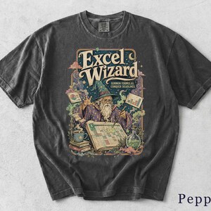May include: A charcoal gray t-shirt featuring a graphic of a wizard in a pointed hat working on a spreadsheet. The text reads Excel Wizard, Summon Formulas, Conquer Deadlines. The design includes charts, books, a coffee mug, and a potion bottle.