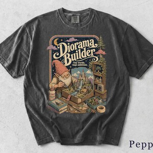 Könnte beinhalten: Ein anthrazitfarbenes Kurzarm-T-Shirt mit einem Grafikdruck eines Gnoms, der ein Miniatur-Burgdiorama baut. Der Text lautet "Diorama Builder" und "Tiny worlds, huge ambitions." Die Illustration zeigt Bücher, Kleber und Werkzeuge in einer warmen Farbpalette.