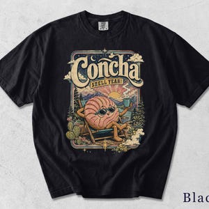 May include: A black short-sleeve t-shirt featuring a graphic print of a pink concha bread character wearing sunglasses, relaxing in a beach chair with a mug. The text reads Concha and SHELL YEAH! above a landscape with mountains, trees, and a sun.