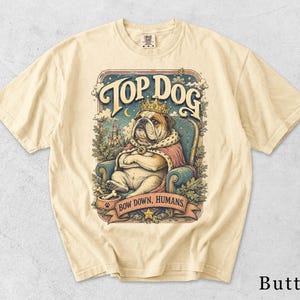Könnte beinhalten: Ein butterfarbenes Kurzarm-T-Shirt mit einer Grafik eines Bulldogs, der eine Krone und einen Umhang trägt und auf einem Stuhl sitzt. Der Text lautet TOP DOG und BOW DOWN, HUMANS. Die Illustration zeigt ein Schloss, Bäume und einen Napf.