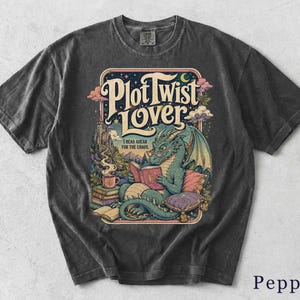 Könnte beinhalten: Ein anthrazitfarbenes T-Shirt mit einem Grafikdruck eines grünen Drachen, der eine Brille trägt und ein Buch liest. Der Text lautet Plot Twist Lover und I read ahead for the chaos. Die Illustration zeigt ein Schloss, eine dampfende Tasse und Bücher.