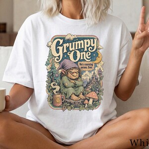 Puede incluir: Una camiseta blanca con un estampado gráfico colorido de una criatura gruñona con gorro morado sentada sobre un tocón. El texto dice Grumpy One y Not a morning person. Ever. La ilustración incluye una taza con un sol, setas, árboles y un castillo.
