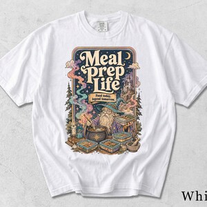 Könnte beinhalten: Ein weißes T-Shirt mit einer farbenfrohen Grafik eines Zauberers, der in einem Kessel rührt, umgeben von Essensbehältern. Der Text lautet Meal prep Life und Food today, legend tomorrow. Das Design zeigt ein Schloss, Bäume und Rauch.