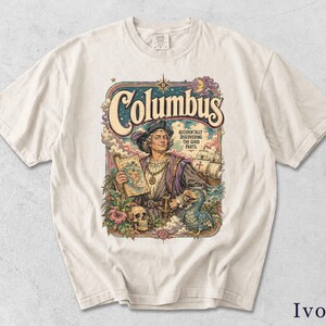 Könnte beinhalten: Ein elfenbeinfarbenes T-Shirt mit einem farbenfrohen Grafikdruck einer historischen Figur, die eine Karte hält, mit dem Text 'Columbus' oben und 'ACCIDENTALLY DISCOVERING THE GOOD PARTS' rechts, umgeben von einem Dodo, einem Totenkopf und einem Schiff.