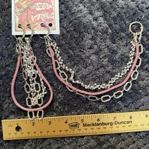 May include: A multi-strand chain accessory featuring pink rhinestone-encrusted cord and silver-tone metal links, attached to a branded card labeled Bootlets and Bearrings by Joelle Joden, measured against a yellow ruler showing 9 inches in length.