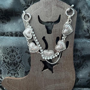 May include: A silver-toned chain necklace featuring multiple embossed heart-shaped charms and delicate white faux pearl accents. The jewelry piece is displayed on a brown wooden boot-shaped stand with a cutout bull head and star design.