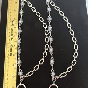 May include: Two silver-toned metal chains featuring oval-shaped white stone inlays with grey veining, connected to circular rings and lobster clasps. A yellow ruler is placed alongside, showing the chains measure approximately 11 inches in length.