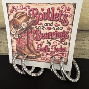 May include: Two pairs of silver-toned hoop earrings featuring embedded clear crystals. The earrings are displayed on a white card with a graphic print of a cowboy hat and pink boots, featuring the text Bootlets and Bearrings by Joelle Jerden.