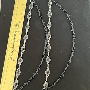 May include: Two silver-toned chains featuring oval black cabochon settings and black barbed wire links, each ending in a circular ring. A yellow ruler is placed alongside, showing the chains measure approximately 11.5 inches in total length.