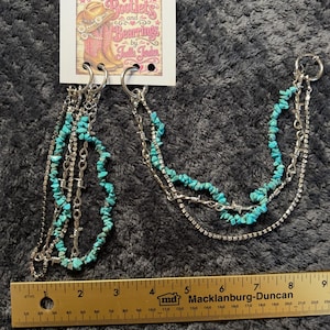 May include: A pair of silver-toned boot chains featuring turquoise chip beads, clear rhinestone strands, and metal barbed wire accents. The chains are attached to a white display card labeled Bootlets and Bearings by Joelle Jorden, measuring 9 inches.