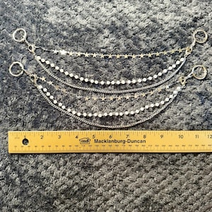 May include: A multi-layered silver-toned chain accessory featuring small star charms and white beads, attached to circular rings at each end. A yellow ruler is positioned below, showing the accessory spans approximately 10.5 inches in length.