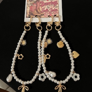 May include: Two beaded lanyards featuring white faux pearls and gold-toned metal charms including suns, flowers, seashells, hearts, and bows. Each lanyard is attached to a gold lobster clasp and displayed on a card labeled Bootlets and Bearings by Joelle Jerden.