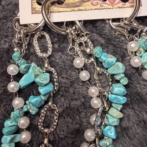 May include: A pair of silver-toned earrings attached to a display card labeled Joelle Tanden. The earrings feature multiple dangling strands composed of polished white pearls, irregular turquoise stone chips, and textured silver-toned metal links.