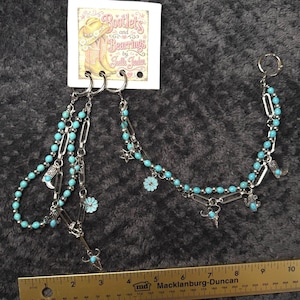May include: A silver-toned chain accessory featuring turquoise-colored beads, small metal charms shaped like cowboy boots, stars, flowers, and skulls, attached to a display card labeled Bootlets and Bearings by Joelle Jorden, with a 10-inch ruler.