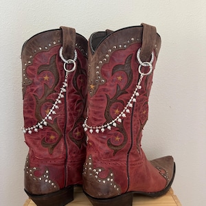 May include: A pair of red and brown leather cowboy boots featuring dark brown overlays, star-shaped cutouts, and silver-tone metal studs. Each boot is adorned with a removable silver-tone chain accessory featuring small white faux pearls and crystals.