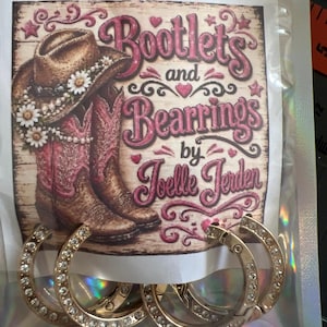 May include: Two pairs of gold-toned hoop earrings encrusted with clear rhinestones, displayed on a branded card featuring a graphic of a cowboy hat and pink western boots with the text Bootlets and Bearrings by Joelle Jerden.