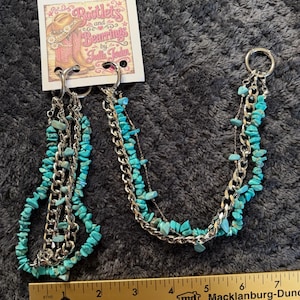 May include: A pair of silver-toned boot chains featuring turquoise-colored stone chips and metal links, attached to a display card labeled Bootlets and Bearings by Joelle Jeulen, positioned above a ruler measuring approximately 7.5 inches in length.