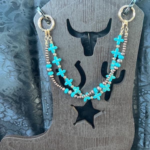 May include: A double-strand beaded necklace featuring bright turquoise cross-shaped beads interspersed with small metallic copper-toned spheres. The necklace is attached to two gold-toned circular clasps and is displayed on a dark brown boot-shaped stand.