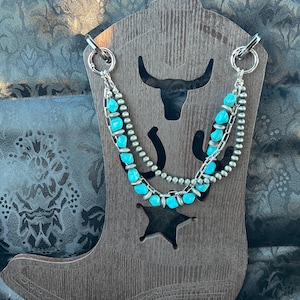 May include: A multi-strand necklace featuring bright turquoise-colored beads and metallic silver-toned beads on a silver chain. The necklace is displayed on a dark brown wooden stand shaped like a cowboy boot with cutouts of a bull and a star.
