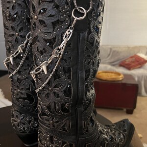 May include: A pair of black knee-high boots featuring intricate floral cutouts, shimmering sequins, and small silver studs. Each boot is adorned with a detachable silver-toned metal chain accessory that includes dangling spikes and a circular ring.