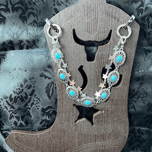 May include: A silver-toned chain necklace featuring oval-shaped concho links with embedded turquoise-colored stones, interspersed with small silver-toned star charms. The necklace is displayed on a brown wooden cutout shaped like a cowboy boot.
