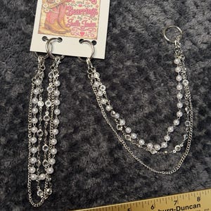 May include: A silver-toned boot chain accessory featuring multiple strands of white faux pearls and clear crystals attached to metal rings. The product is displayed on a branded card labeled Bootlets and Bearrings by Joelle Jerden, positioned above a ruler measuring 8 inches.