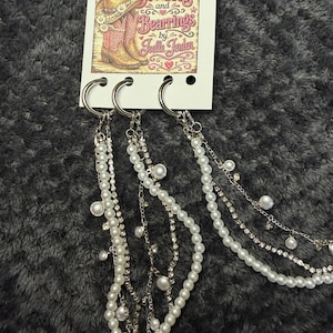 May include: Three silver-toned metal rings attached to a white display card featuring the text Bootlets and Bearrings by Joelle Jerden. Each ring holds multiple dangling chains adorned with white faux pearls and clear rhinestones in various lengths.
