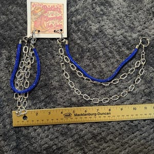 May include: A set of two boot chains featuring blue glitter-textured bands and silver-toned oval link chains with clear rhinestones. The chains are attached to a branded display card and measured against an 11-inch yellow wooden ruler.