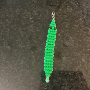 May include: A long, narrow, woven keychain or zipper pull crafted from bright green plastic beads. The design features a repeating pattern with white accent edges and two small black beads near the top, finished with a silver metal lobster clasp.