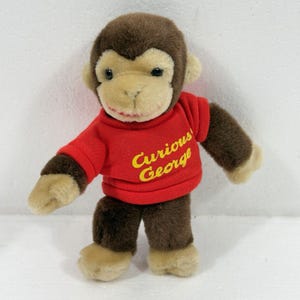 May include: A plush monkey toy with brown and tan fur wearing a red short-sleeved shirt. The shirt features the text Curious George printed in yellow script across the chest. The monkey has dark eyes, a tan face, and tan hands and feet.