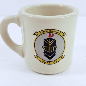 May include: A cream-colored ceramic mug featuring a circular emblem with a dark blue knight helmet graphic, a red plume, and the text Black Knights VMFA 314 printed in black on a yellow banner surrounding the central gray circle design.