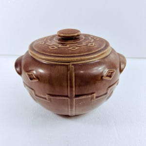 May include: A round, brown ceramic lidded jar with a glossy finish. The exterior features raised geometric patterns, including diamond shapes and linear cross designs. It has two small side handles and a matching circular lid with a central knob.