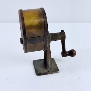 May include: A vintage manual pencil sharpener featuring a cylindrical brass body with a dark metal frame and base. The device includes a side-mounted hand crank with a small wooden handle and a metal plate embossed with the number 2 on the side.