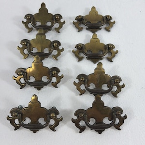 May include: A set of eight brass drawer pulls arranged in two columns. Each pull features a decorative backplate with a central fleur-de-lis shape and ornate scrolling edges, paired with a matching curved swing handle attached by two small knobs.