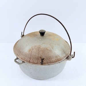 May include: A round, silver-colored metal pot with a matching domed lid, featuring a dark, thin wire bail handle and a small black knob on top. The exterior surface shows signs of wear, including rust-colored discoloration and a textured finish.